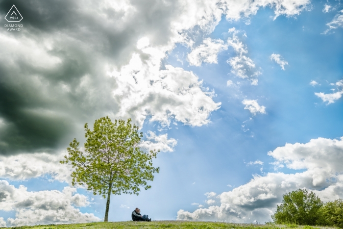 Engagement photo session of a couple sitting on their hill next to a tree beneath a sunny, blue sky in Saint Martin du Touch, France.