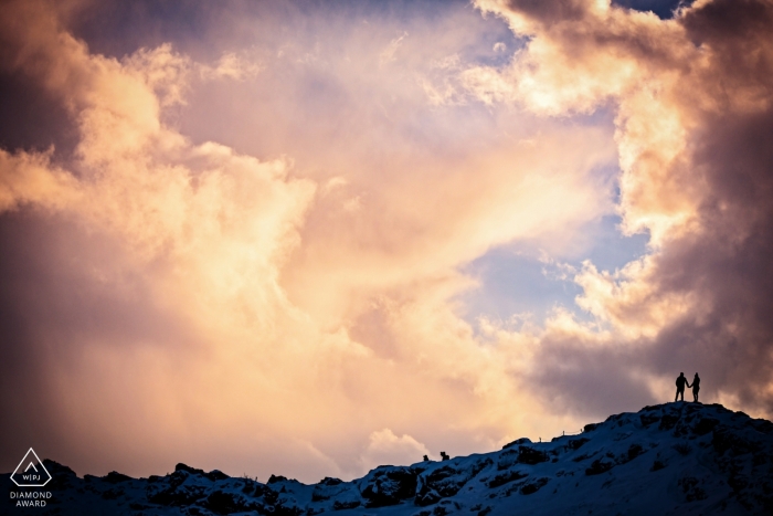 Foto do acoplamento de um par que está sobre um monte nevado no nascer do sol cercado por nuvens de tempestade em Islândia.