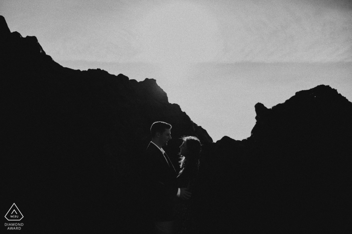 Séance de fiançailles noir et blanc d'un couple debout devant une grande formation rocheuse à Bodega Bay, Californie.