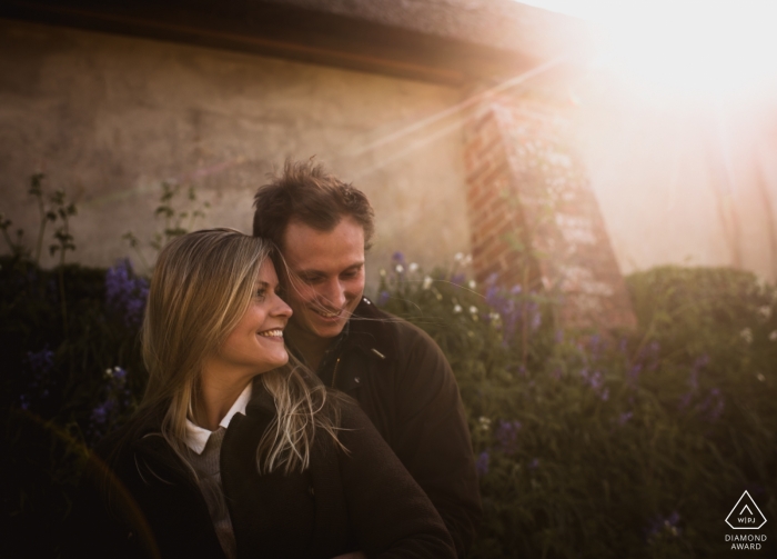 Foto de compromiso de una pareja de pie frente a una casa de piedra y flores mientras el sol brilla en Broadchalke en Wiltshire, Inglaterra.