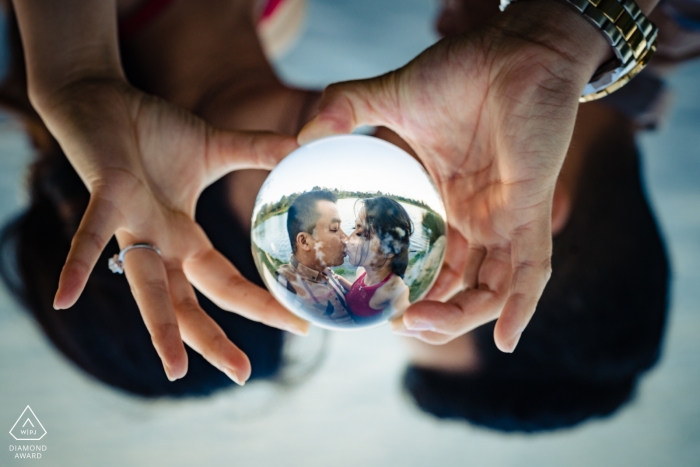Photo by: <a href="https://www.wpja.com/photos/diamond-award-top-wedding-engagement-photographer/2464791">Leo Do - Fullerton California Engagement portrait of a couple kissing reflected in a glass ball they are holding together in El Monte, CA.