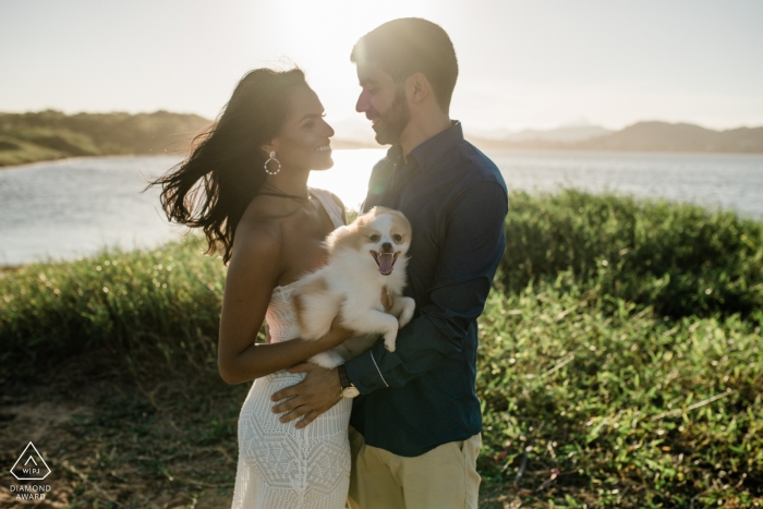 Macae engagement photo shoot shows the couple smiling happily at each other while holding their dog Macae engagement photo shoot shows the couple smiling happily at each other while holding their dog