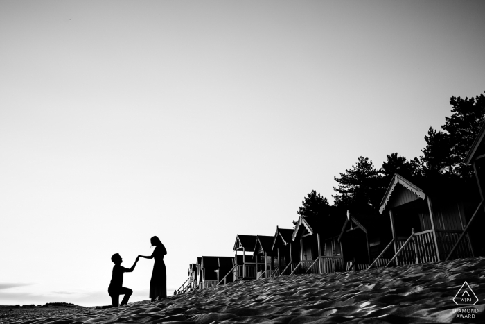 Wells-next-the-Sea, Royaume-Uni - Un homme pose sur un genou à côté de sa future épouse alors qu'une rangée de cabanes borde la plage lors de cette séance de photographie de fiançailles en noir et blanc