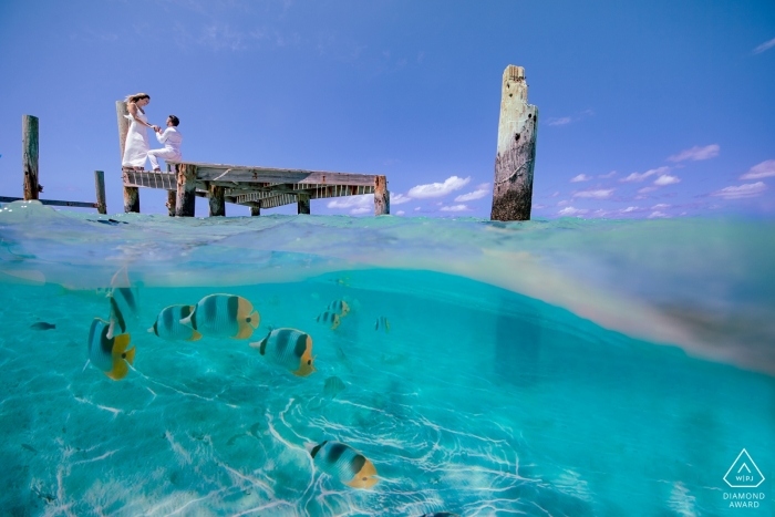 Bei diesem Verlobungsfotoshooting auf den Bahamas wird ein Paar, das auf dem Dock posiert, von einem Schwarm bunter Fische, die sich direkt unter den Wellen verstecken, fotografiert