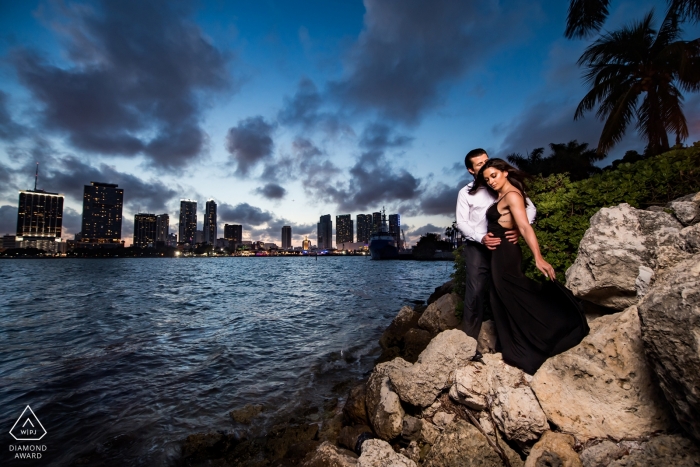 Pareja posa en las rocas junto al océano, mientras que el horizonte de Miami está lejos en esta sesión de fotos de compromiso