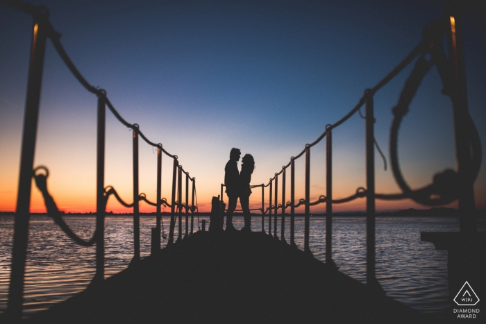 Photo by: <a href="https://www.wpja.com/photos/diamond-award-top-wedding-engagement-photographer/2472141">Marco Vegni - Siena Siena Orbetello, Tuscany engagement portrait shoot - couples silhouettes are captured as they stand on a long pier in the ocean as the sun sets behind them