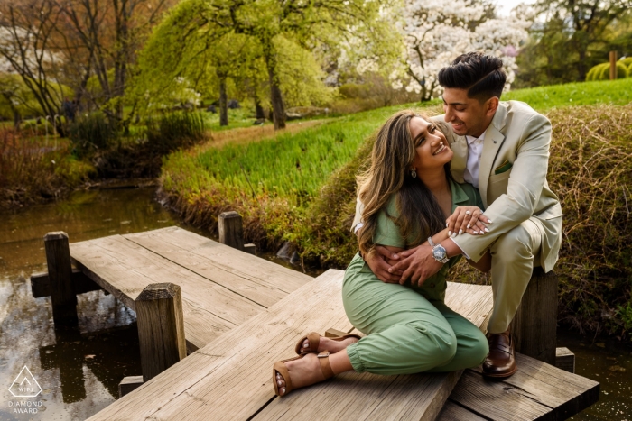 A couple smiles at each other while sitting on a dock during their engagement shoot at Montreal Botanical Gardens