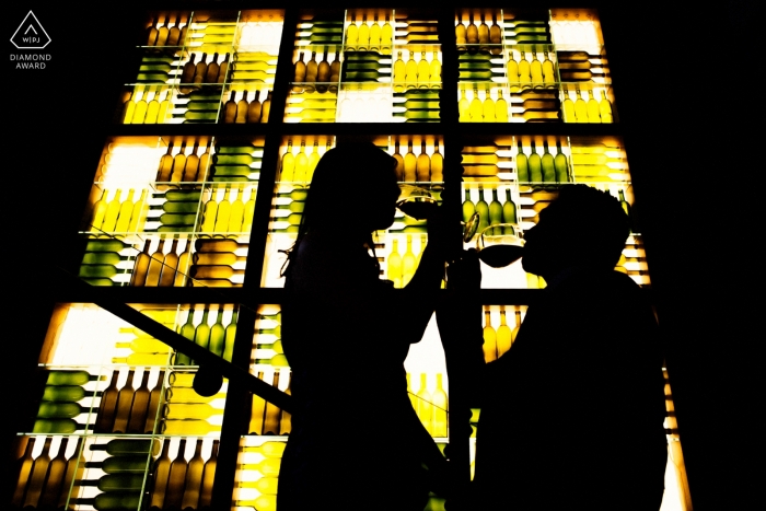 The silhouettes of the couple sipping wine in front of a green and brown stained glass window was created during a San Francisco engagement photo shoot 