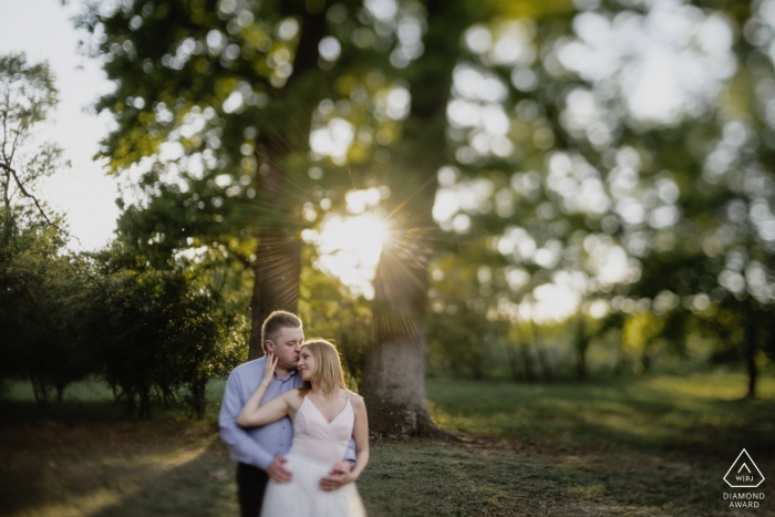 Photo by: <a href="https://www.wpja.com/photos/diamond-award-top-wedding-engagement-photographer/2475516">Krisztian Bozso - Budapest Budapest Csengele, Hungary - a man kisses his future bride on her forehead as sun shines through the trees in this engagement photo session