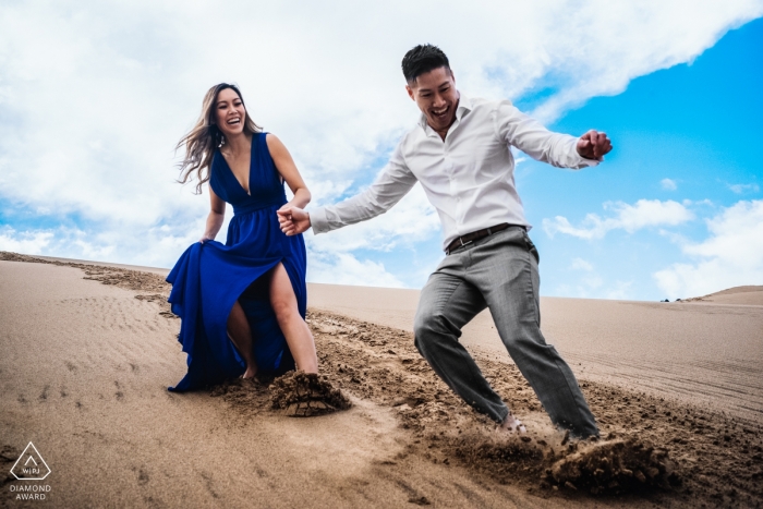 Foto por: <a href="https://www.wpja.com/photos/diamond-award-top-wedding-engagement-photographer/2476486"> Dorota Karpowicz - Edmonton Alberta Fotografía de compromiso del Valle de la Muerte: una pareja que se ríe se desliza descalzo por las dunas de arena bajo un cielo azul brillante