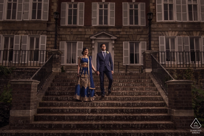 Photo by: <a href="https://www.wpja.com/photos/diamond-award-top-wedding-engagement-photographer/2476541">Jay Hoque - Uxbridge London London, UK engagement photo session. Couple stands holding hands on stone steps leading up to a large gray building