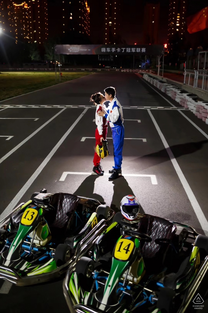 A couple kisses in racing gear on a race track at night in this engagement photo session by a Shandong, China photographer.