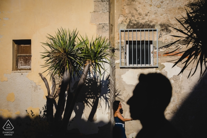 A woman stands holding the large shadow of a man's chin on Orgigia Island during their engagement photoshoot by a Syracuse, Sicily photographer.