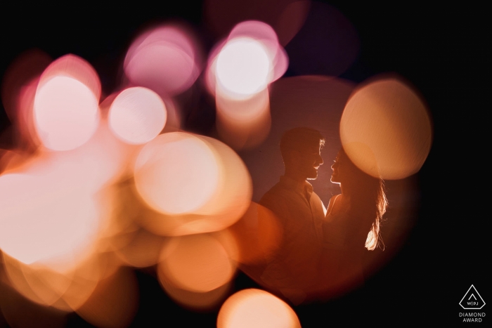 The couple can be seen standing on the other side of a circular window as lights reflect around them during their engagement session by a London, England photographer