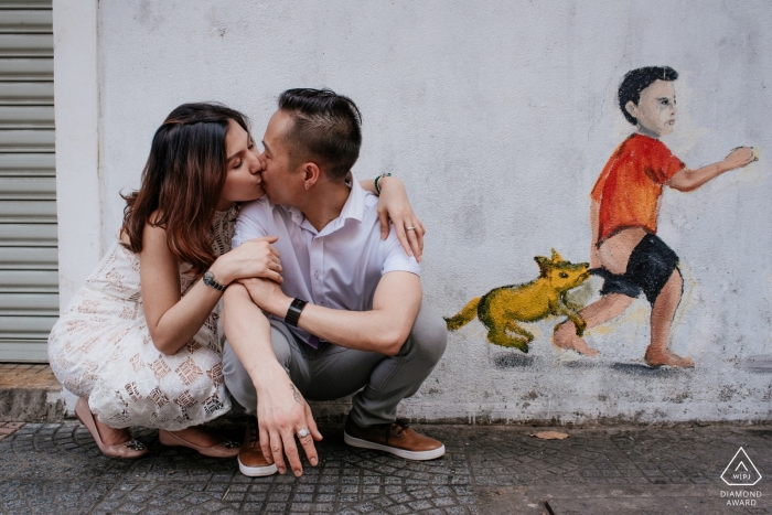 A couple squats on the ground kissing in front of a wall in Ho Chi Minh with graffiti art of a boy and a dog in this engagement photo by a Vietnam photographer.