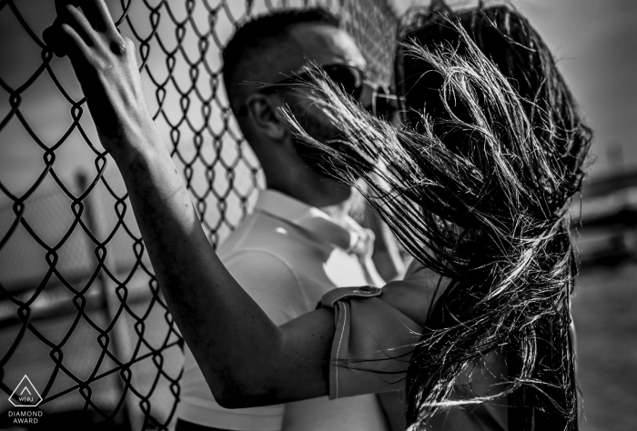 A woman's hair blows in the wind in Cabo de Gata as she and her fiance stand against a fence in this black and white pre-wedding photo by a Murcia, Spain photographer.