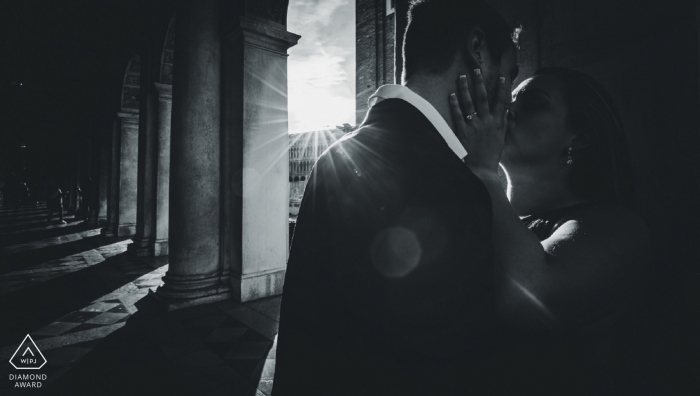 Photo by: <a href="https://www.wpja.com/photos/diamond-award-top-wedding-engagement-photographer/2485311">Carlo Bettuolo - Venezia Venezia A couple kisses as the sun beams through large columns in this black and white pre-wedding portrait by a Venice, Veneto photographer.