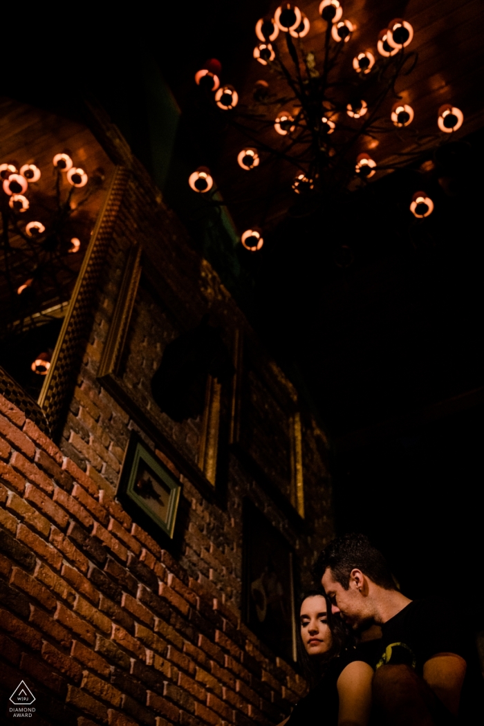 Un couple se tient à côté d'un mur de briques sous un lustre à Rio das Ostras pour sa séance photo précédant le mariage par un photographe de Rio de Janeiro.