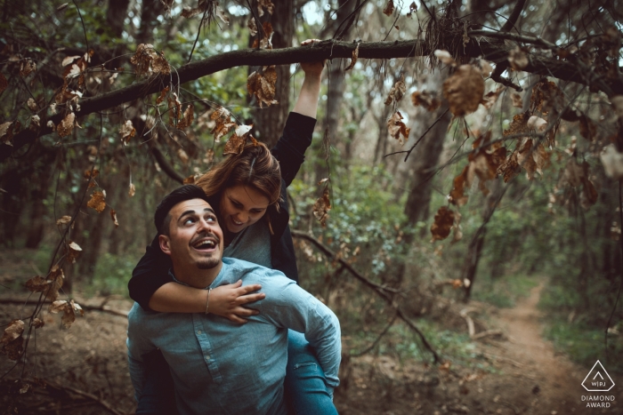 Foto von: <a href="https://www.wpja.com/photos/diamond-award-top-wedding-engagement-photographer/2485471"> Tyler Nardone - Scauri Latina Ein Mann trägt seinen Verlobten auf dem Rücken, während sie sich in dieser Verlobungssitzung eines Latina-Fotografen an einem Ast im Wald von San Felice Circeo festhält.