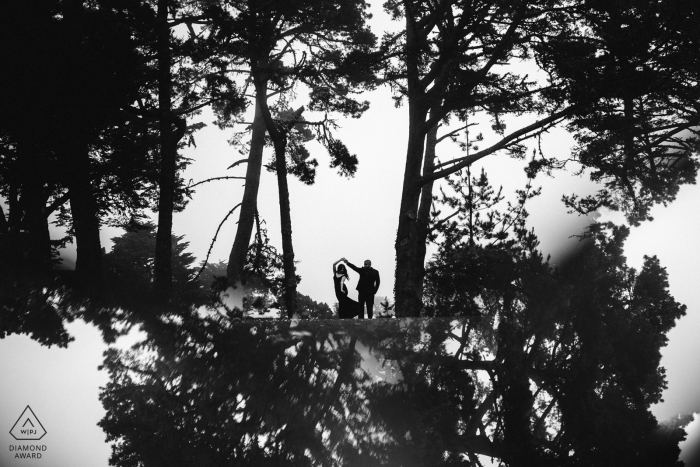 A man twirls a woman as they stand among tall trees in San Francisco in this black and white engagement photo by a Sacramento, CA photographer.