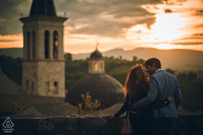 Un couple se tient devant une tour de Spoleto et s’embrasse au coucher du soleil lors de la séance de photos de fiançailles de Perugia, photographe de l’Ombrie.