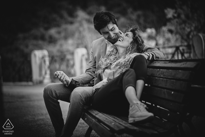 Em Spoleto, um homem e uma mulher sentam-se alegremente juntos num banco e riem-se nesta foto preto e branco pré-nupcial feita por um fotógrafo da Perugia, Umbria.