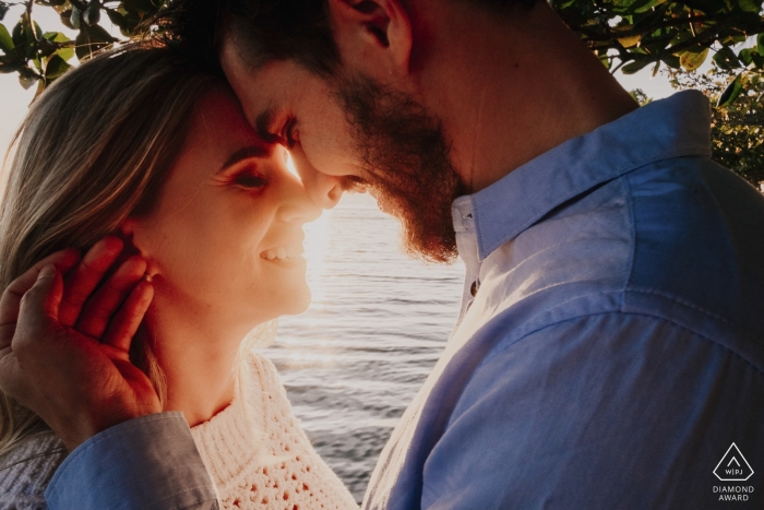 A man and woman hold their heads together as sun shines between them during their Laguna engagement session by a Santa Catarina, Brazil photographer.