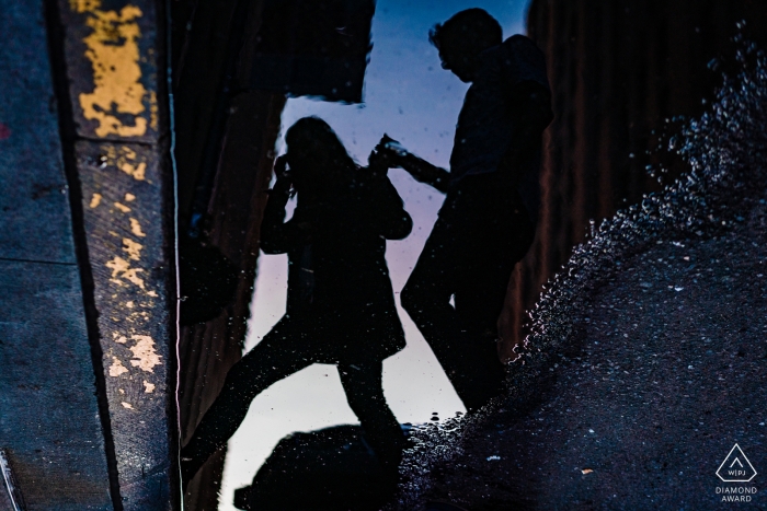 A man can be seen helping a woman cross the street in the reflection of a puddle during their pre-wedding photo session by a New York City photographer.