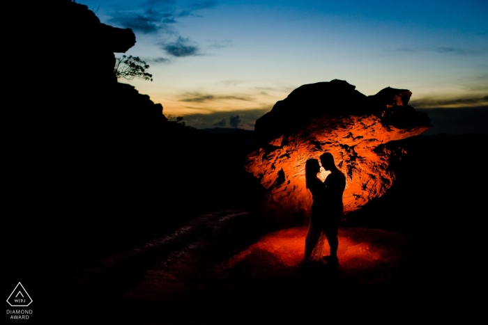 A couple in Lavras Novas is silhouetted as they stand together in front of a large rock formation at dusk in this pre-wedding photo session by a Minas Gerais, Brazil photographer.