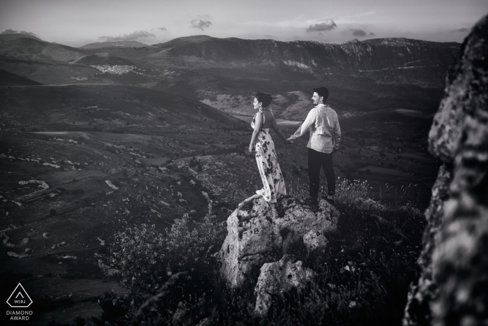 Una pareja se toma de la mano mientras se paran en una roca en Rocca Calascio con vistas a las llanuras en esta sesión de compromiso de un fotógrafo de Lazio, Italia.