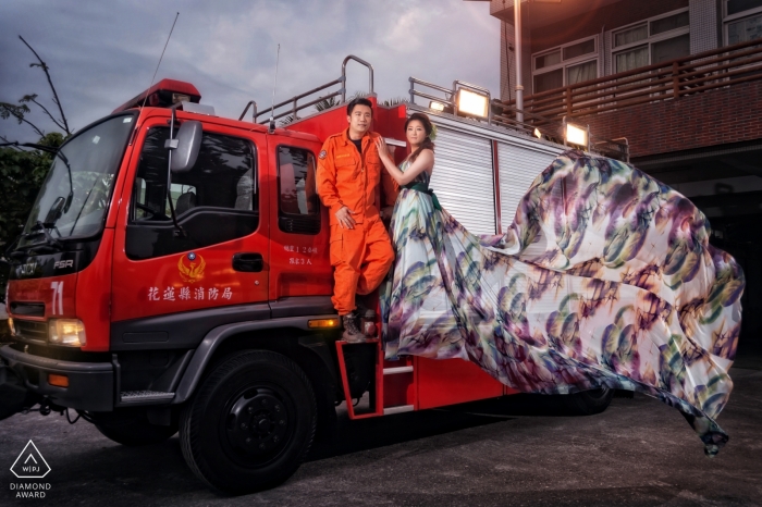 A woman's dress blows dramatically in the wind as she and her fiance stand together on a large, red truck in this engagement portrait by a Hualien County, Taiwan photographer.