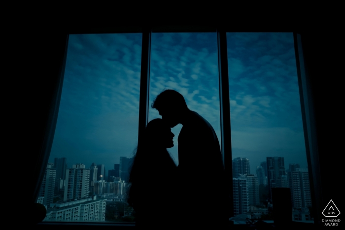 A man kisses his fiance on the forehead as they stand near a window looking out on the night sky of Singapore in this engagement portrait by a Tamil Nadu, India photographer.