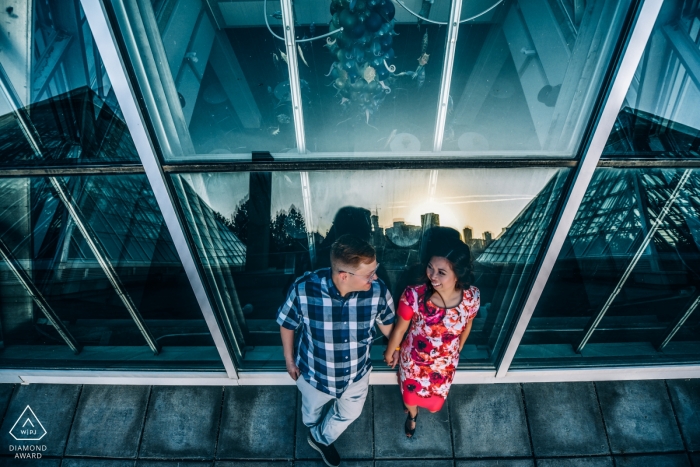 A couple stands against a building in Edmonton in front of tall windows in this engagement photo by an Alberta, Canada photographer.