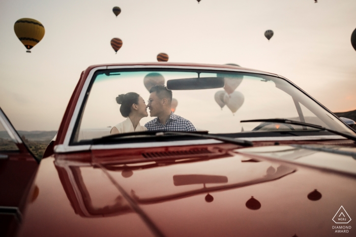 Una pareja comprometida se sienta en el auto y se besa mientras los globos aerostáticos vuelan alrededor de ellos durante su sesión de compromiso en Turquía, Capadocia.