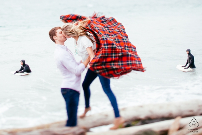 Séance photo avant le mariage à Seattle, Washington - Un homme s'accroche à sa fiancée alors que les surfeurs sont à l'arrière-plan