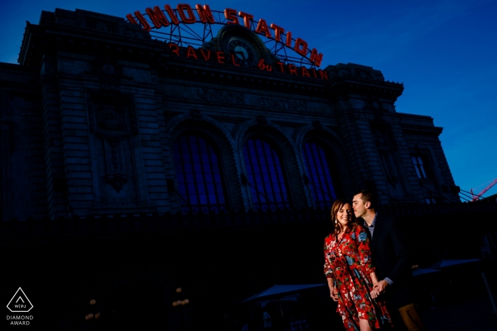 Usando algo de luz disponible para resaltar a la pareja comprometida contra la Union Station en el centro de Denver durante una sesión de retratos.