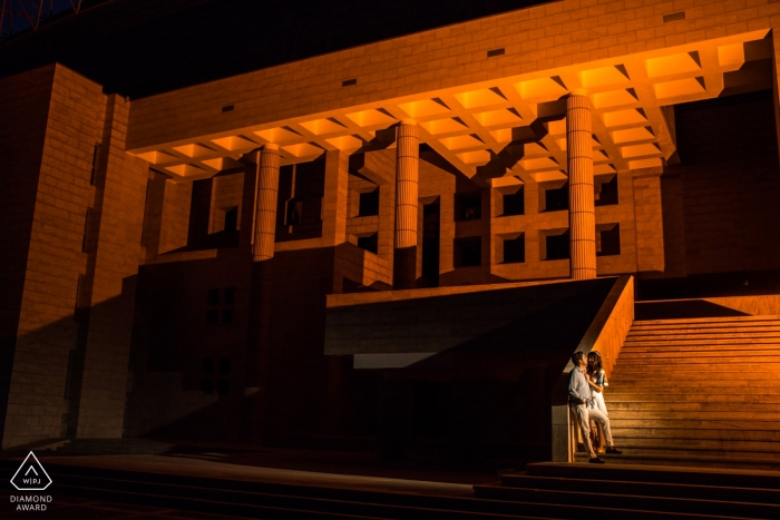 Un couple fiancé se tient devant un amphithéâtre rehaussé de couleur orange au campus principal de l’Université de Bilkent, Ankara