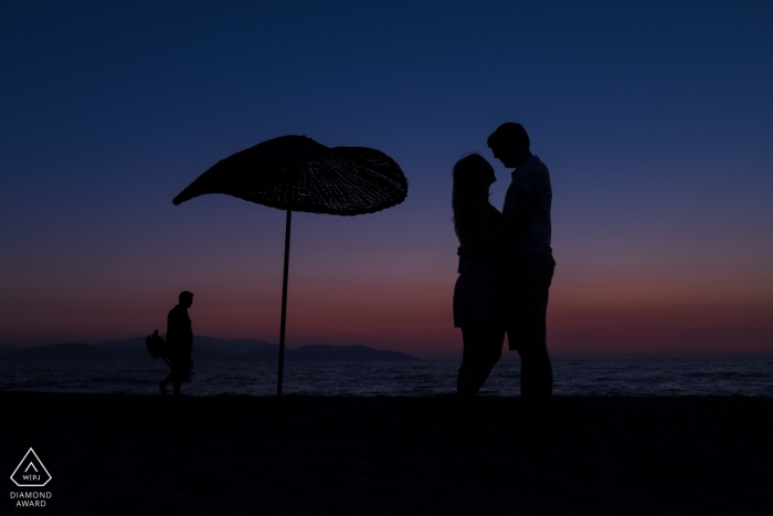 Photo by: <a href="https://www.wpja.com/photos/diamond-award-top-wedding-engagement-photographer/2559973">Orçun Yalçın - Ankara Ankara Güzelçamlı Kuşadası, Turkey Pre-Wedding Portrait Session — Silhouette of the couple and a sun umbrella at the beach with a fisherman passing behind of them