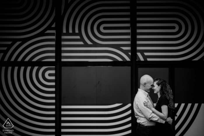 Photo by: <a href="https://www.wpja.com/photos/diamond-award-top-wedding-engagement-photographer/2560125">Brittany Diliberto - Arlington Virginia Crystal City, VA engagement photo session in black and white | A couple laughs together in front of a black and white striped window covering.