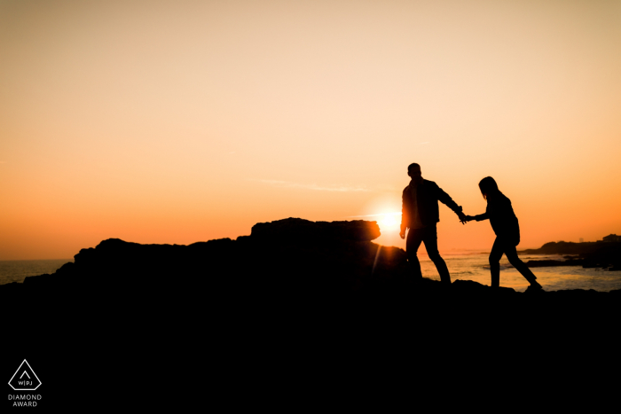 Silhouette picture of a couple walking on rocks on a cliff above the ocean at sunset | Engagement photographer