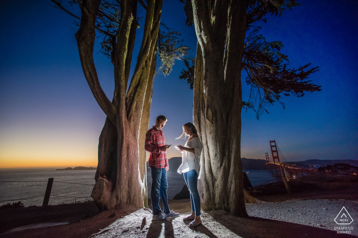 San Francisco, CA Paare auf ihren Handys während eines beleuchteten Verlobungsporträttriebs bei Sonnenuntergang.