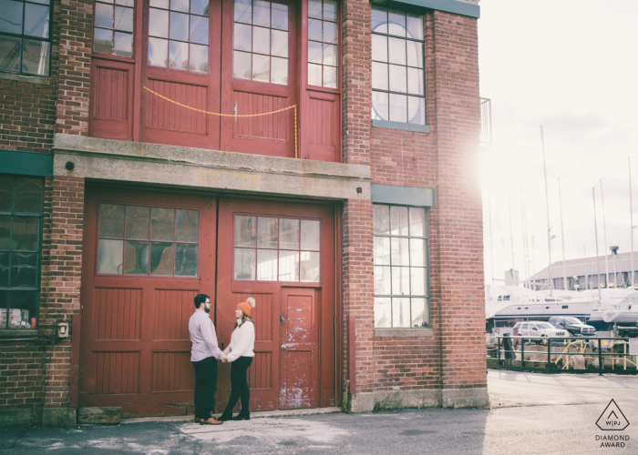 Séance de fiançailles - Le couple se tient devant les bâtiments industriels de l'East Boston Shipyard