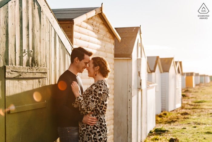 Shoreham Beach, West Sussex, UK pre-wedding portrait session | Couple embracing by the beach huts with sun flare 