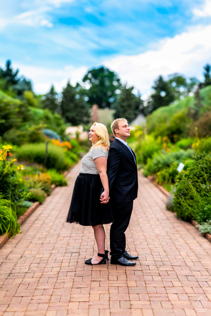 Photographie de fiançailles | Jardins botaniques de Denver, Denver, CO - Un couple engagé se tient dos à dos et admire les jardins - un panorama de Brenizer