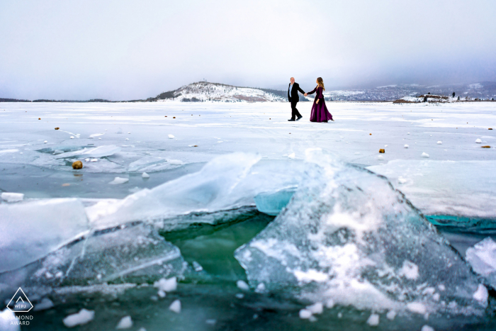 Ein Paar geht Hand in Hand, während es während seiner Verlobungssitzung an einem windigen Wintertag in der Nähe von Silverthorne, Colorado, am zugefrorenen Lake Dillon entlang spaziert.