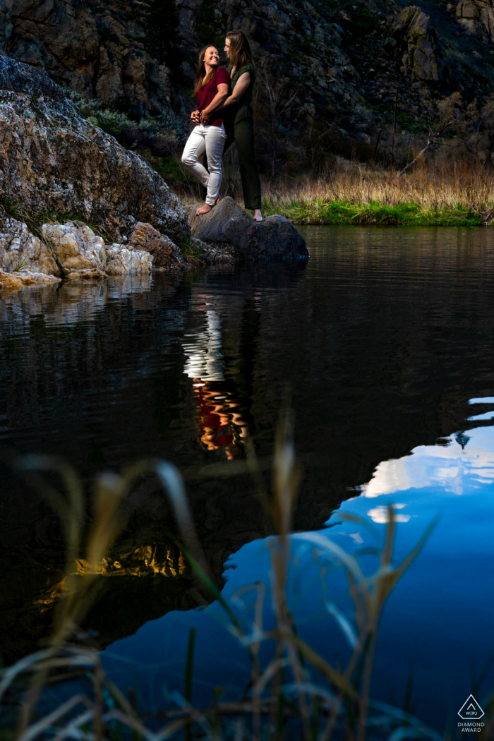 A couple shares a laugh as they pose during their engagement session with reflections on the water A couple shares a laugh as they pose during their engagement session at Gateway Natural Area in the Poudre Canyon near Fort Collins, Colorado.