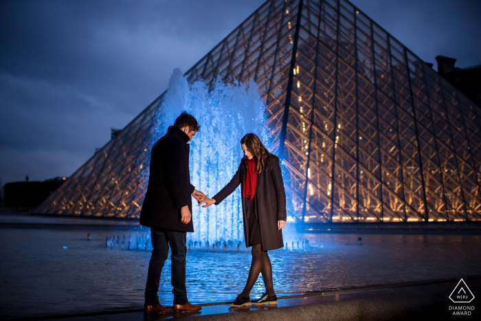 Sessão de fotos de noivado no pirâmide do Louvre de um casal com uma fonte de água brilhando atrás deles.