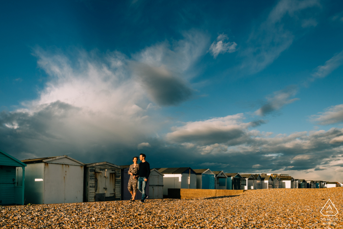 Couple walking along beach huts under dramatic stormy skies at Shoreham Beach, West Sussex, UK 	 