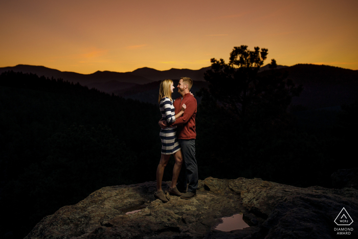 O sol se põe atrás de um casal no topo do Mirante do Irmão durante suas fotos de noivado no Alderfer Three Sister's Park em Evergreen, Colorado