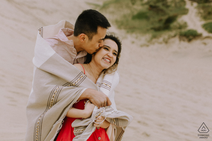 Un couple en riant sur la plage enveloppé dans une couverture lors d'une séance de portrait de fiançailles à Cape Kiwanda, Oregon